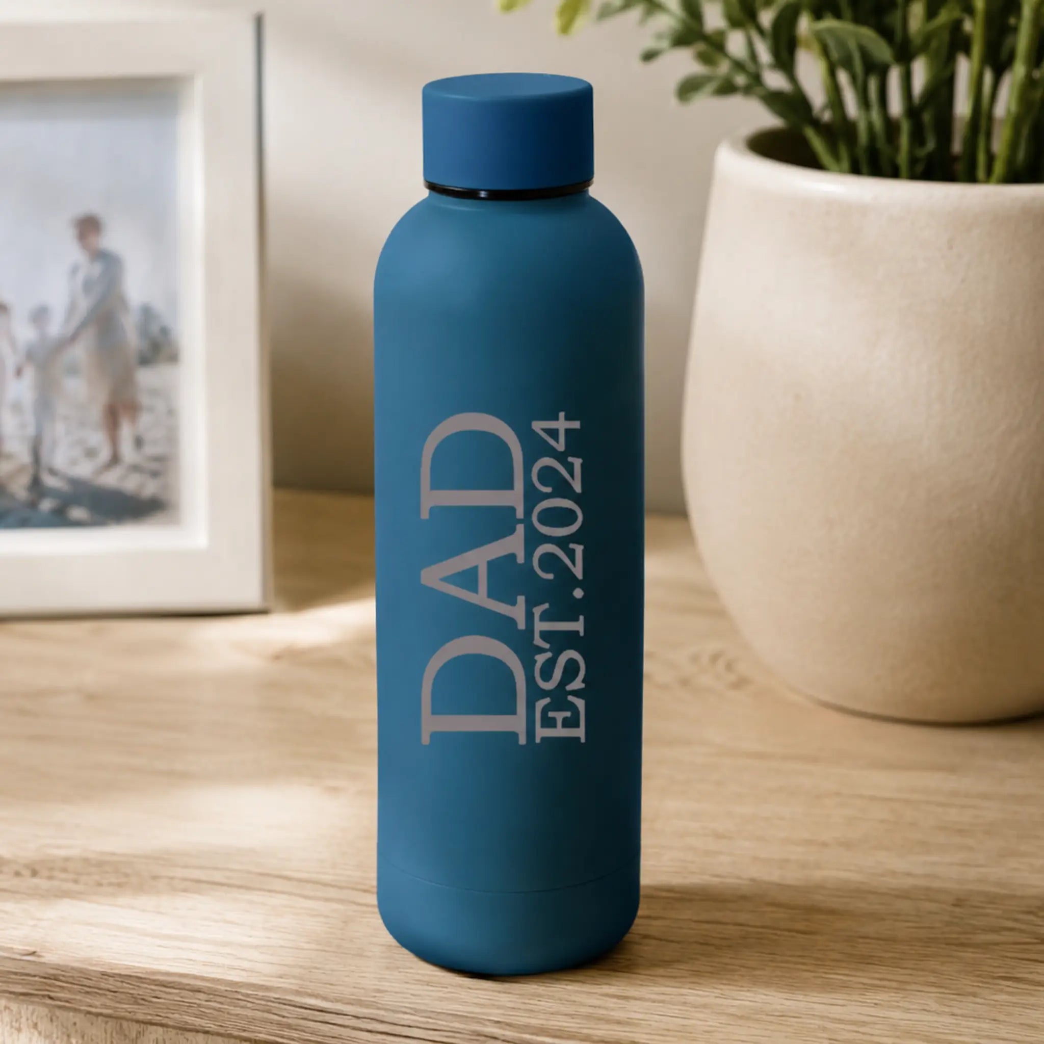 Personalised blue insulated water bottle with “DAD EST. 2024” printed in white text, displayed on a wooden table beside a plant and framed family photo in a warm home setting.
