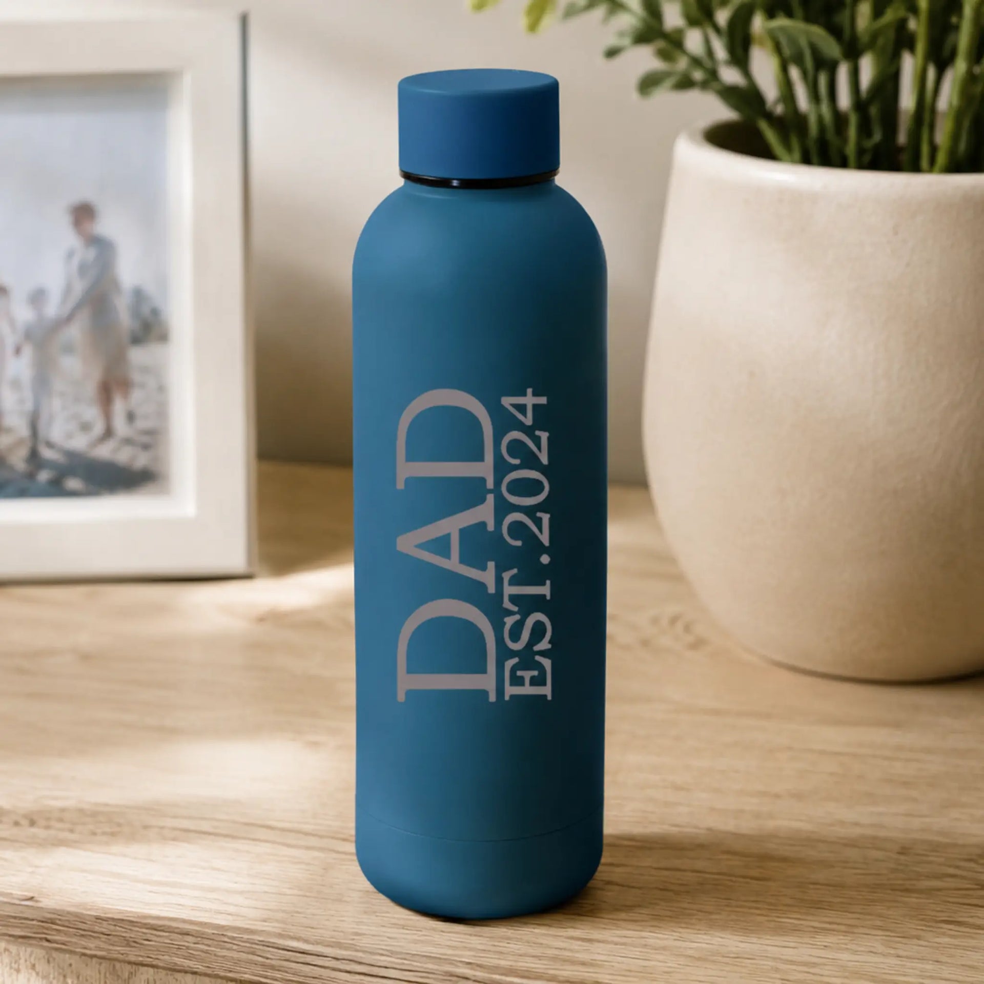 Personalised blue insulated water bottle with “DAD EST. 2024” printed in white text, displayed on a wooden table beside a plant and framed family photo in a warm home setting.